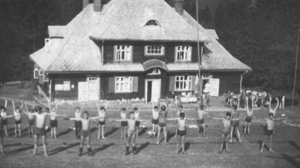 zdjęcie czarno-białe przedstawiające grupę dzieci ćwiczących gimnastykę na zewnątrz, w tle widoczny budynek ośrodka Kordon